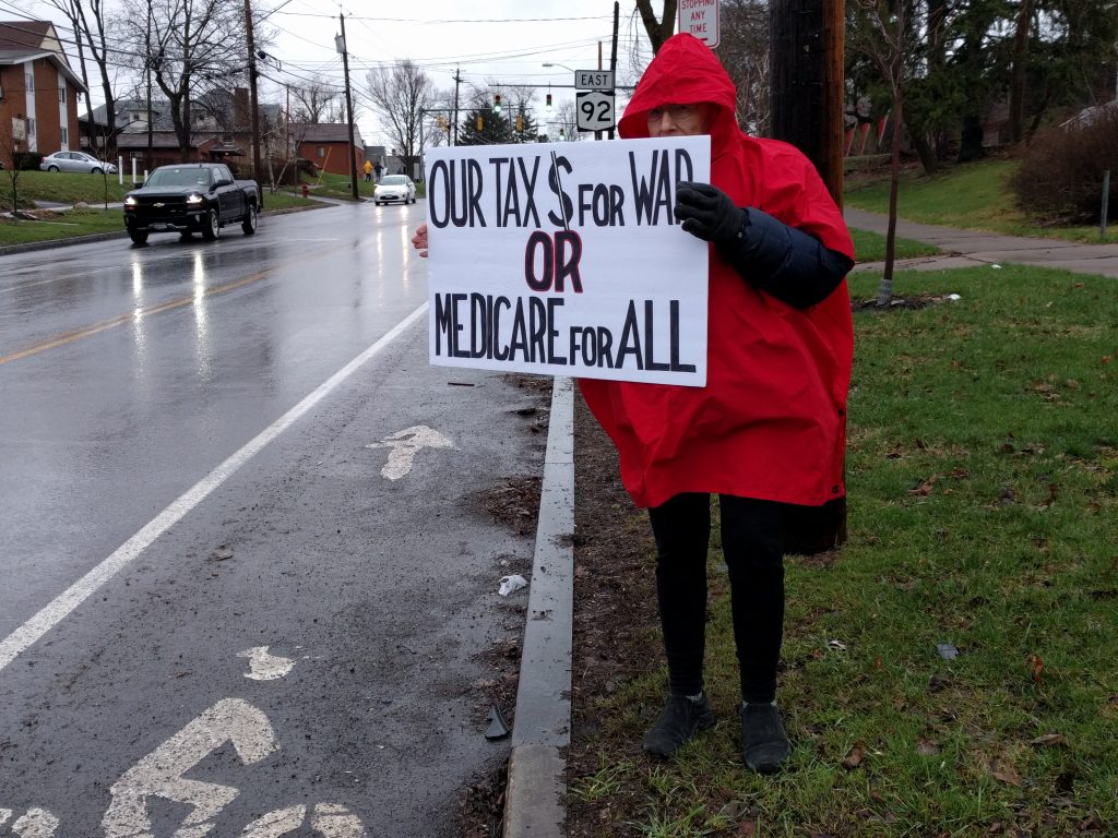 Rae protesting in the rain: Our tax $ for war or Medicare for All.